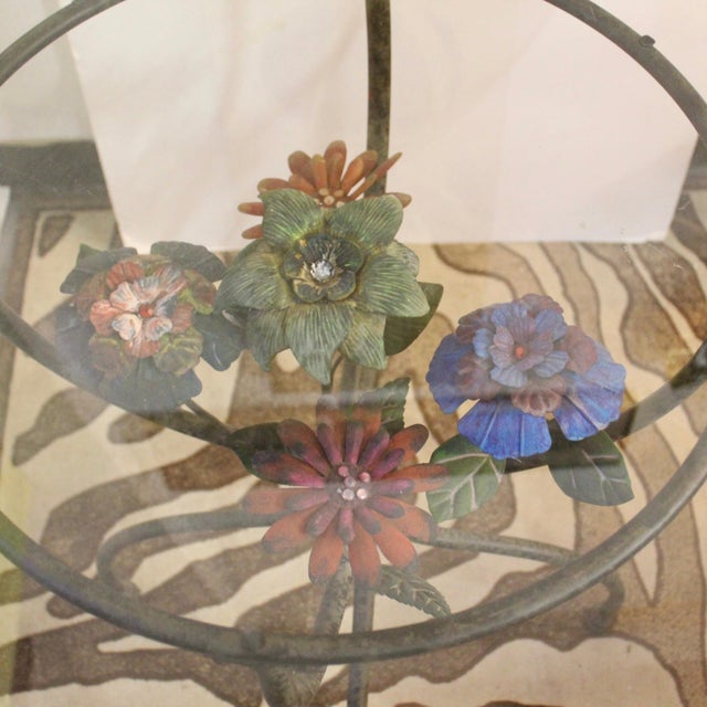 This wrought iron accent table showcases a metal colorful bouquet positioned at the center. Glass top shown is NOT included.