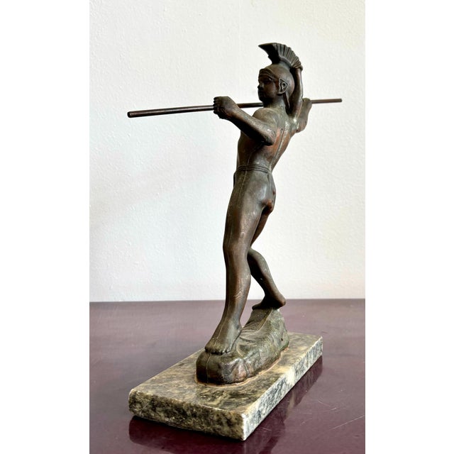 This bronze-toned figure captures the poised intensity of a classical warrior, frozen in the moment before launching his...