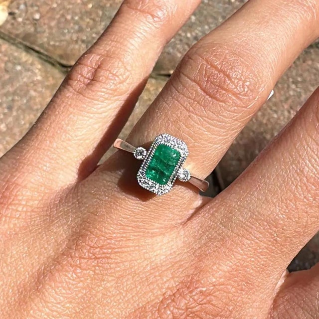 An emerald rests in a rich green at the centre of this solid 14K white gold ring, surrounded by twelve earth-mined...