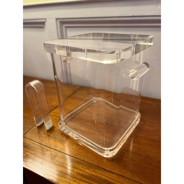Midcentury thick lucite ice bucket with solid cylinder handles, tongs and hinged lid. The bucket has a rounded square...