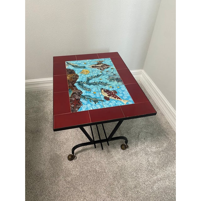Charming French mid-century side table and magazine rack featuring a hand-decorated ceramic tile top with a colorful...