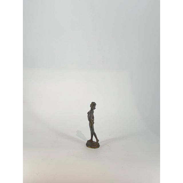 This charming bronze figurine captures a young woman in a moment of quiet curiosity, her hands gently clasped behind her...