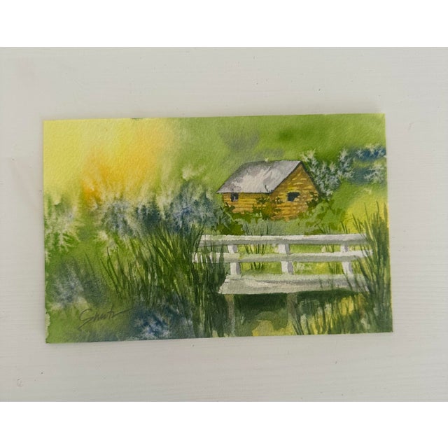 Bathed in soft golden light, this intimate watercolor depicts a small yellow cabin tucked behind tall grasses and a white...