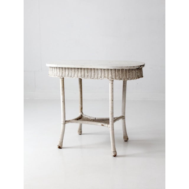 This early 20th-century white wicker table features a softly worn painted finish, an oval wood plank top, and layered...