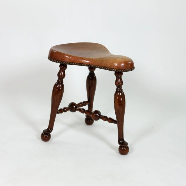 A turned mahogany three-legged saddle-seat stool with a molded leather seat and nailhead trim; on turned legs, joined by...