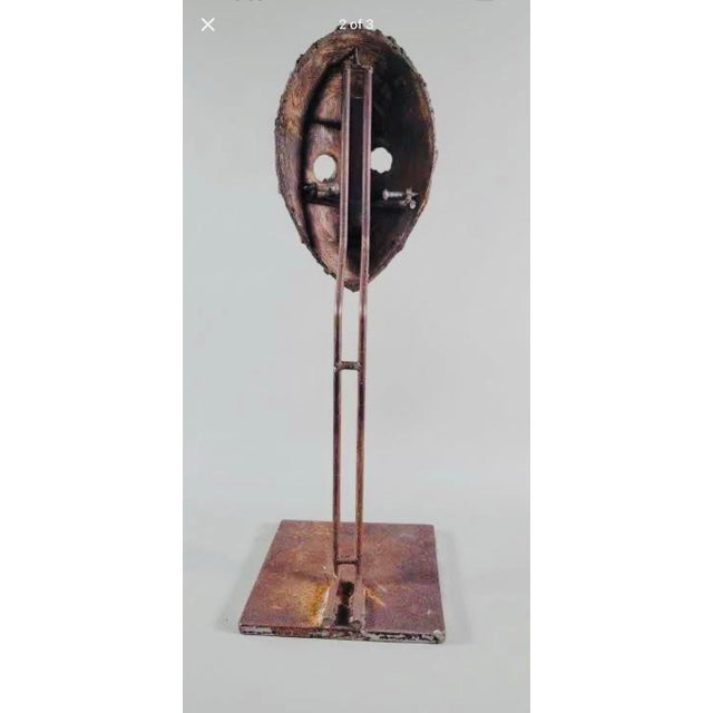 Rustic wood face mask sculpture has a heavily textured surface with brown, black, and red tones. The mask is mounted on a...