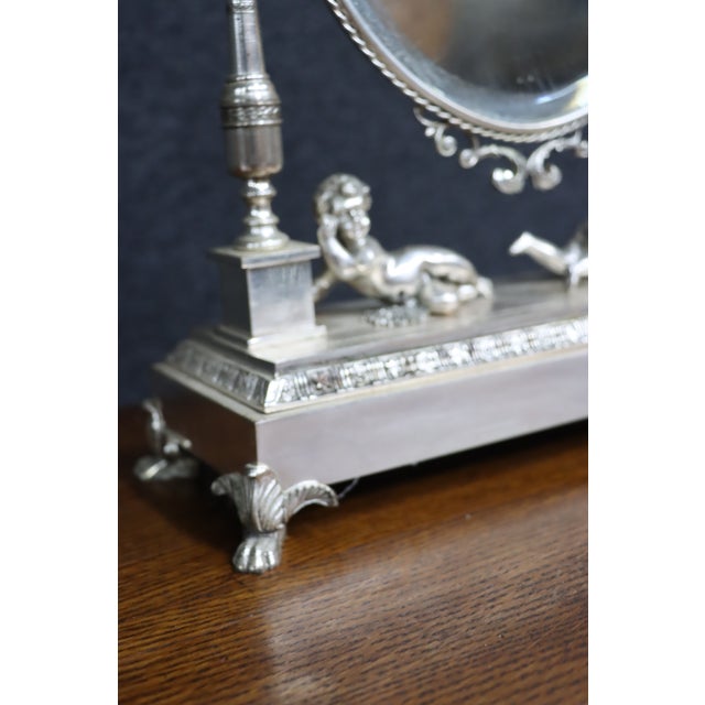 Ornate oval tabletop vanity mirror featuring an elaborate silver-tone frame with scrolling openwork, a decorative crest...