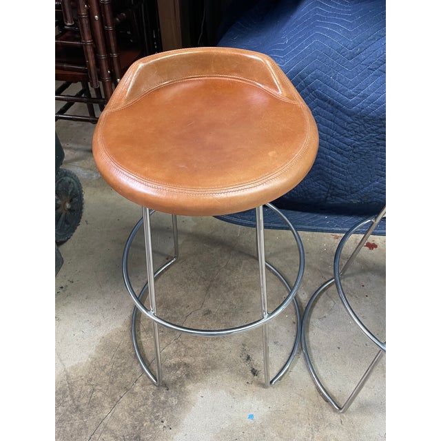 Set of four McGuire swivel counter stools featuring saddle-brown leather seats atop polished stainless steel frames. The...
