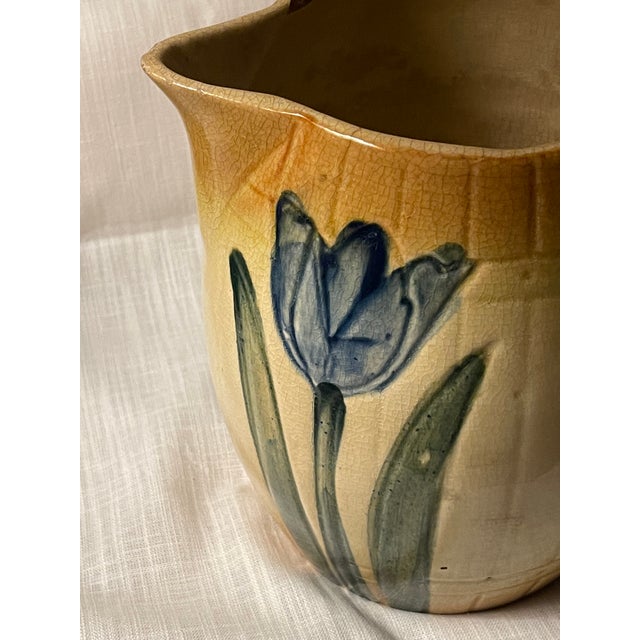 This antique Roseville 1910s ceramic pitcher adorned with bright blue tulips is a beautifully crafted piece of American-...