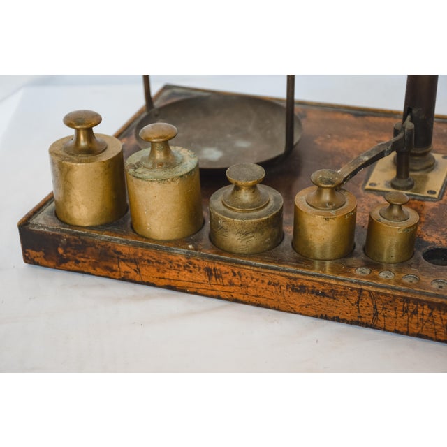 This equal arm brass scale is set on a wooden platform with brass weights. The wooden platform has circular cutouts for...