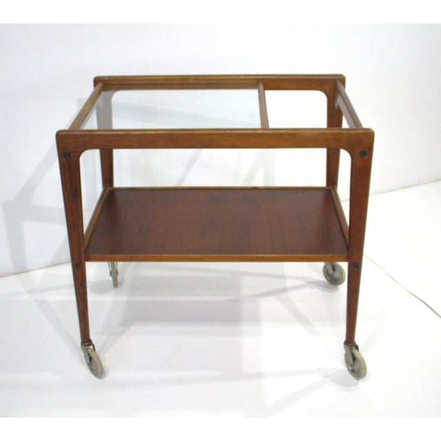 A teakwood framed rolling bar cart with glass inserted top , one end open to be used as a handle and a lower teak storage...