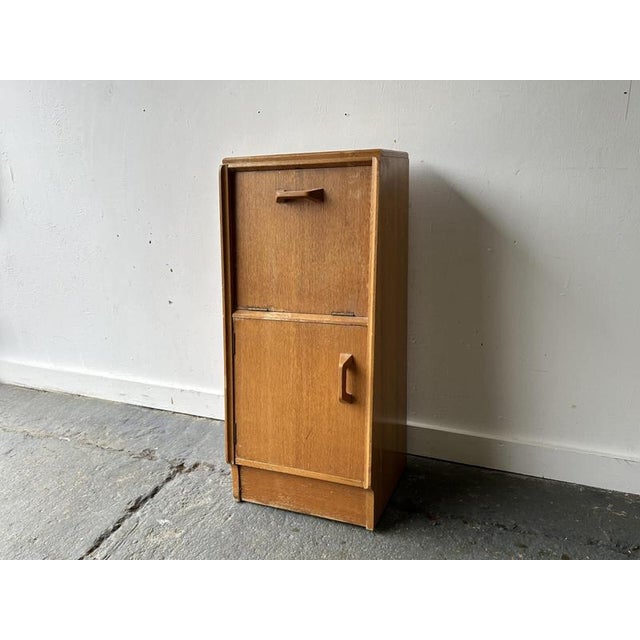 A compact mid-century bedside unit in light oak, featuring a top drop-down cupboard door and a lower hinged cupboard. Part...