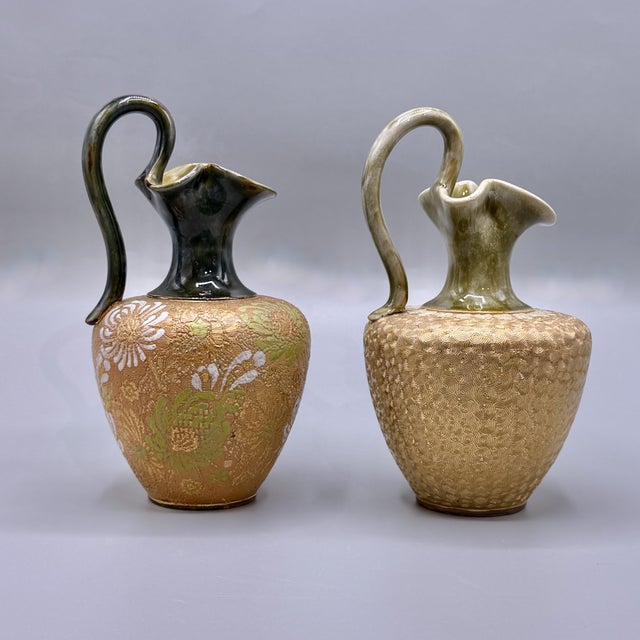A complementary pair of royal doulton lambeth stoneware ewers or handled jugs. standing at a versatile 20 cm in height,...