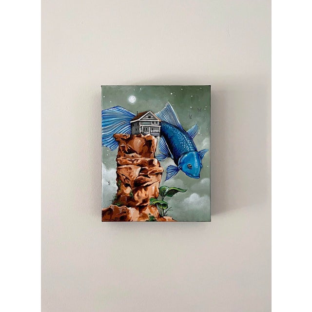 This artwork presents a surreal scene where a large blue fish carries a house atop a rocky outcrop, set against a muted...