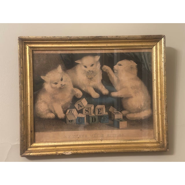 A charming antique Currier & Ives print titled “My Three White Kitties,” depicting three fluffy white kittens playfully...