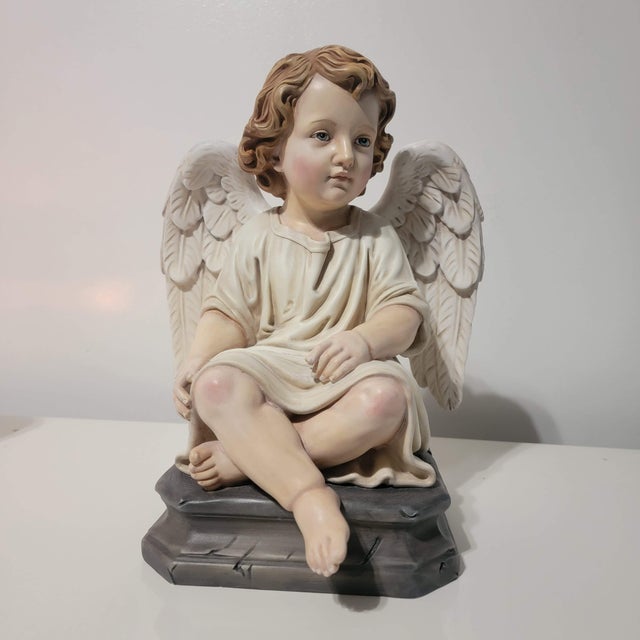 A seated child angel rests on a dark pedestal, with folded legs, hands gathered at the knees and softly feathered wings....