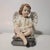 A seated child angel rests on a dark pedestal, with folded legs, hands gathered at the knees and softly feathered wings....