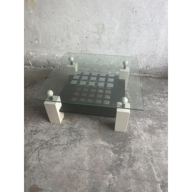 A bold postmodern coffee table featuring a thick square glass top supported by four geometric block legs topped with...