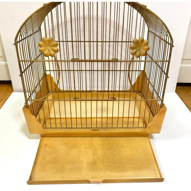 A striking 1950s Hollywood Regency bird cage featuring gold metal bars, floral rosette accents, and a sculptural arched...