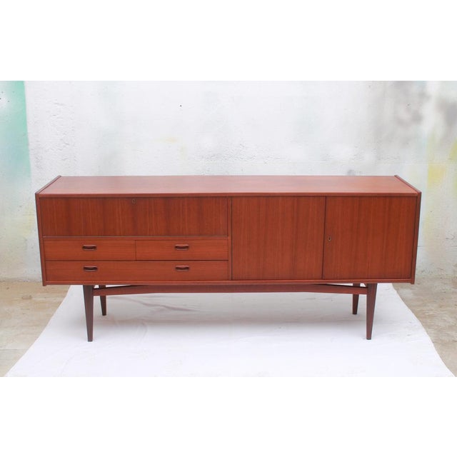 Elegant 1960s sideboard with teak veneered surfaces and a solid teak base. On the left is a drop-down door with one long...