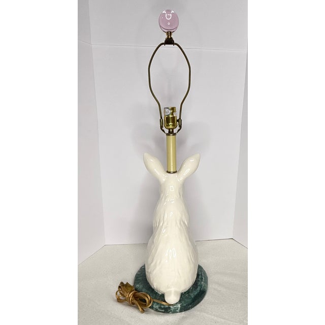 Tall dignified rabbit table lamp features standing rabbit on two feet. White rabbit with pink ears and nose is perched on...