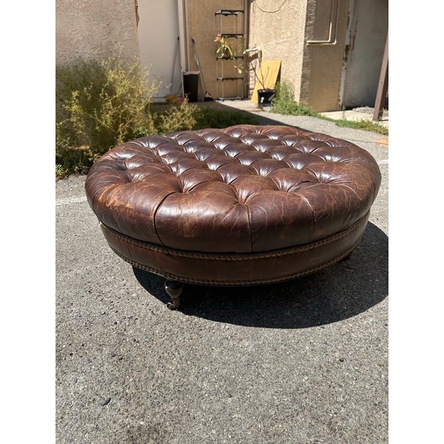 Traditional Chesterfield-style round deep-buttoned tufted leather ottoman, featuring a rich brown distressed leather...