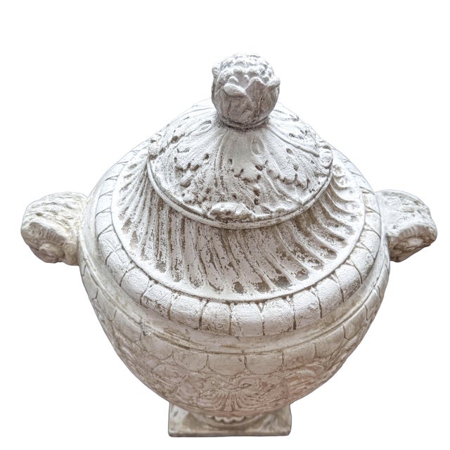 A substantial neoclassical style lidded urn featuring classical ram head handles, fluted body detailing, and an aged...