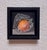 This artwork presents an orange partially enveloped in translucent plastic, creating a striking contrast between the...