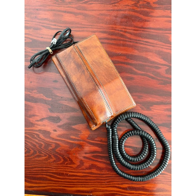 Corded desk telephone covered in stitched tan leather. The slim handset rests on a matching leather base. The keypad sits...