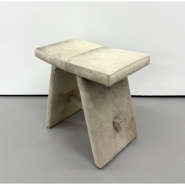 A sculptural stool fully upholstered in natural cream and taupe hair-on-hide, featuring a broad rectangular seat with a...