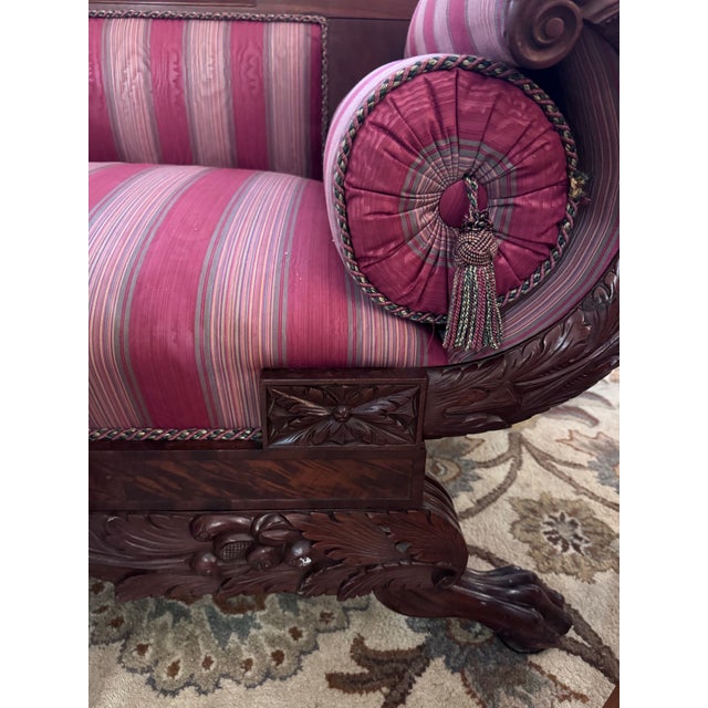 A striking American Empire Revival sofa featuring dramatic rolled scroll arms, richly carved mahogany frame, and...