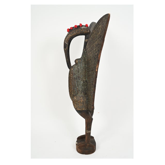 A tall carved wooden West African Senufo hornbill bird sculpture with a tuft of red fabric crowning the head, slender...