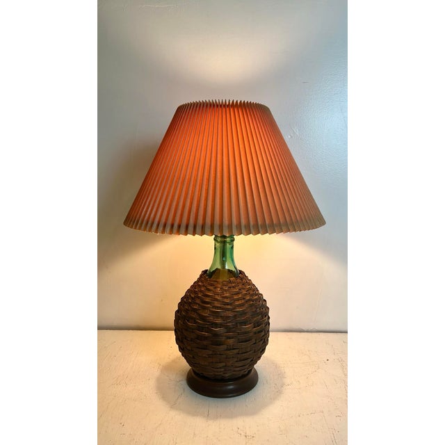 Mid century basket wrapped bottle lamp with accordion shade. Thick woven dark brown reeds around green glass body. Mounted...