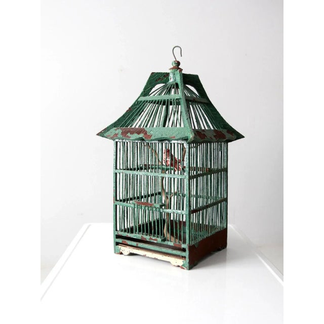 This antique painted wooden birdcage features a charming peaked roof and a gently timeworn finish that enhances its...