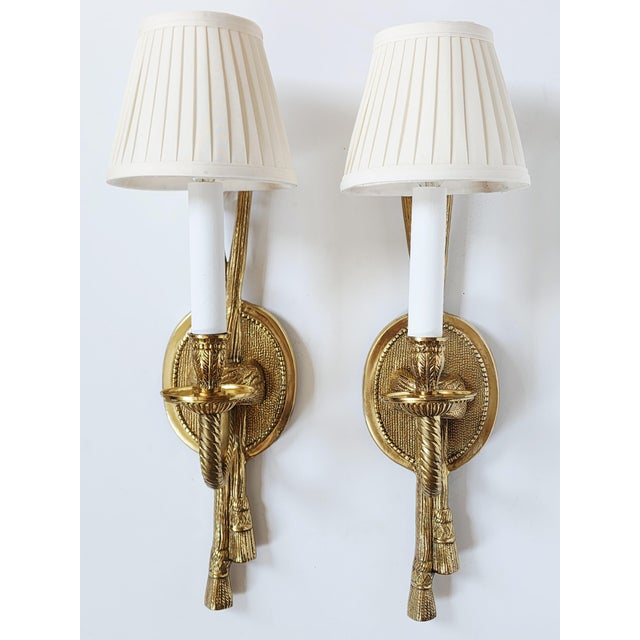 Pair of French Neoclassical style brass wall sconces with one light and silk pleated shades. The sconces feature a tassel...
