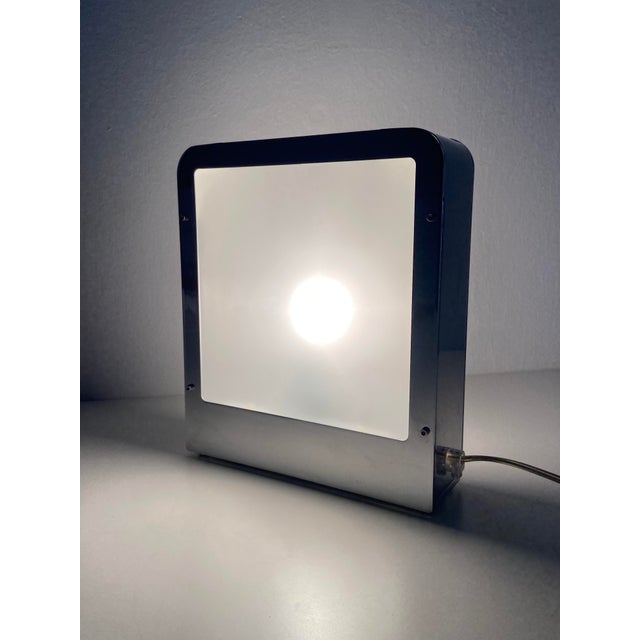 A table lamp with a minimalist, postmodern design, featuring a polished steel frame and a large, double-sided frosted...
