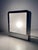 A table lamp with a minimalist, postmodern design, featuring a polished steel frame and a large, double-sided frosted...