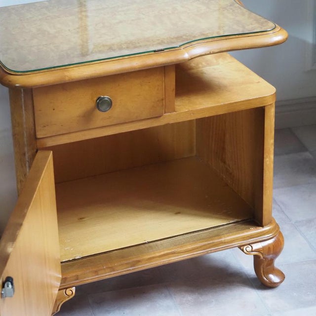A Louis XV-style bedside table, crafted around 1950 as a mid-20th-century period piece. The curved contour of the tabletop...