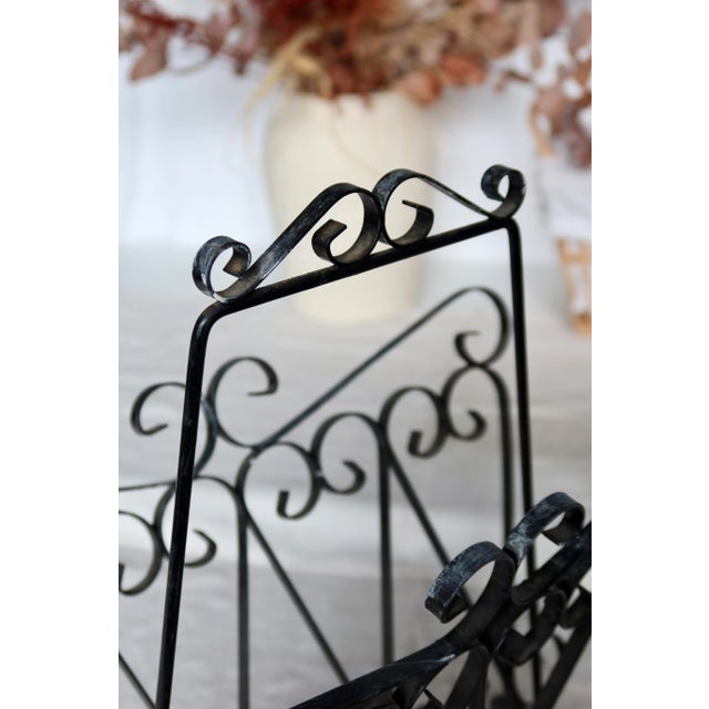 Elegant magazine rack in black lacquered metal, decorated with pretty wrought iron-style scrollwork. It features a central...