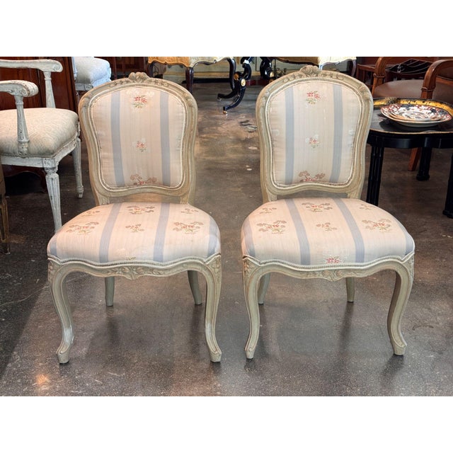 A small pair of French side chairs in an old pale green painted finish, each with a shaped cartouche back crested by a...