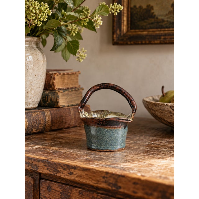 A vintage handmade studio pottery basket featuring a sculptural arched handle, softly flared rim, and rounded stoneware...