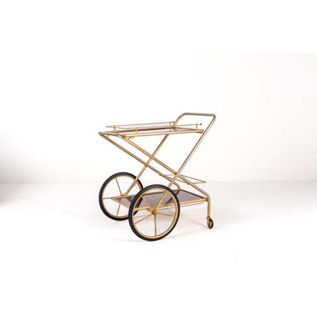 Elegant mid-century folding bar cart featuring a brass-plated tubular frame and large spoked wheels with rubber rims. The...