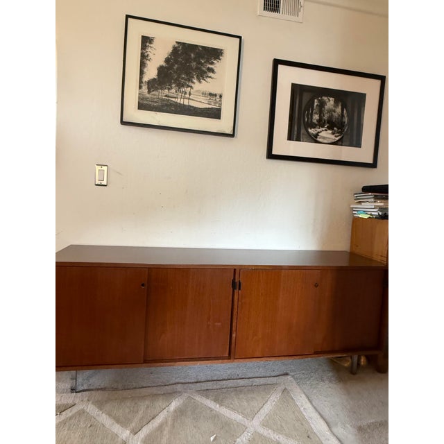 The item is a low wooden credenza featuring a rectangular top, four sliding cabinet doors with small hardware pulls, and a...