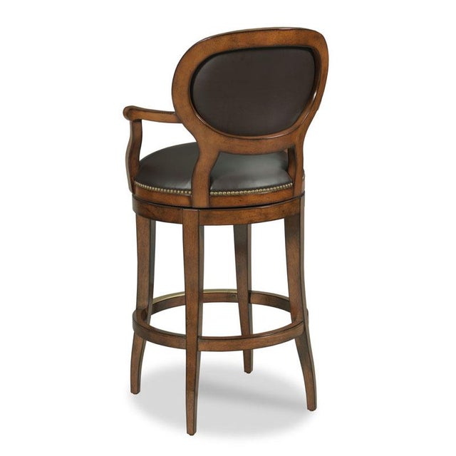 A graceful bar stool built for comfort. An upholstered oval back accented with brass nails is situated above an...