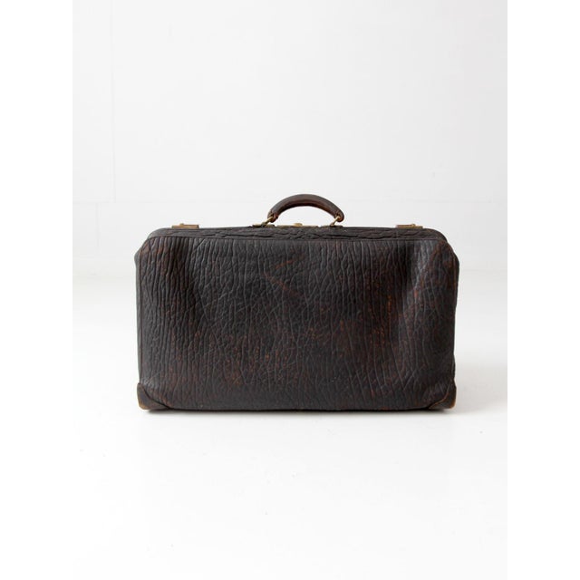An antique black leather suitcase with a substantial form, heavily grained leather, aged brass hardware, and a leather-...