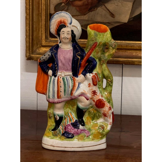 Figure in a plumed hat, purple jacket and striped kilt stands beside a green tree trunk spill with a dog alongside. Lively...