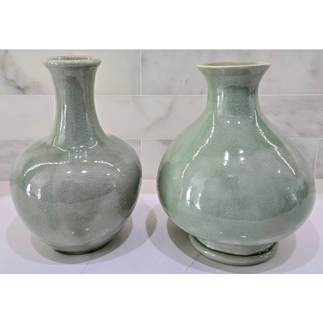 A vintage pair of Thai celadon ceramic vases featuring a soft sage-green finish, glossy crackle glaze, and two distinct,...