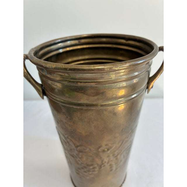 Vintage solid brass Hosley vase with a raised grapevine motif, rolled rim, and curved side handles. The warm, varied...