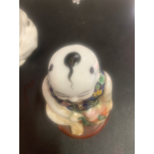 Porcelain figurine in sitting position. Garbed in an embroidered tunic hand painted with flowers and leaves, a small pile...