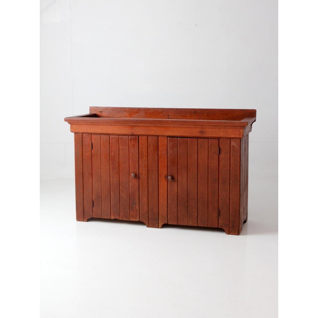A late 19th-century antique wood dry sink with a deep inset top above a two-door cabinet, featuring vertical paneled...